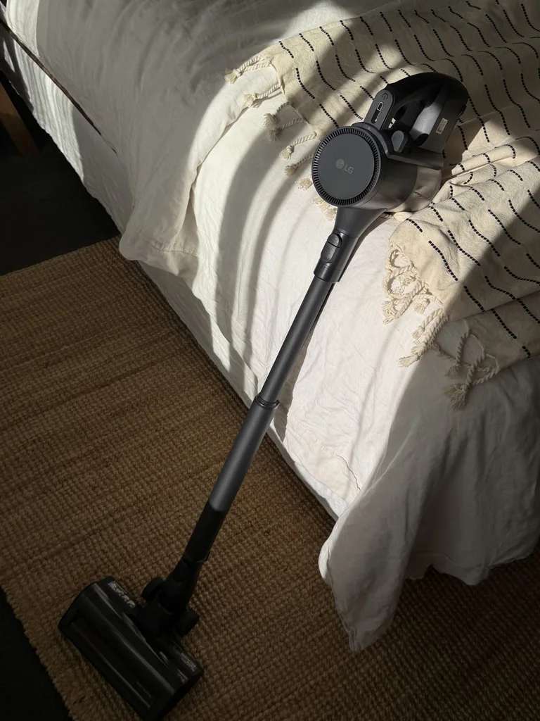 Cordless LG vacuum cleaner leaning against a bed with a woven rug in the foreground.