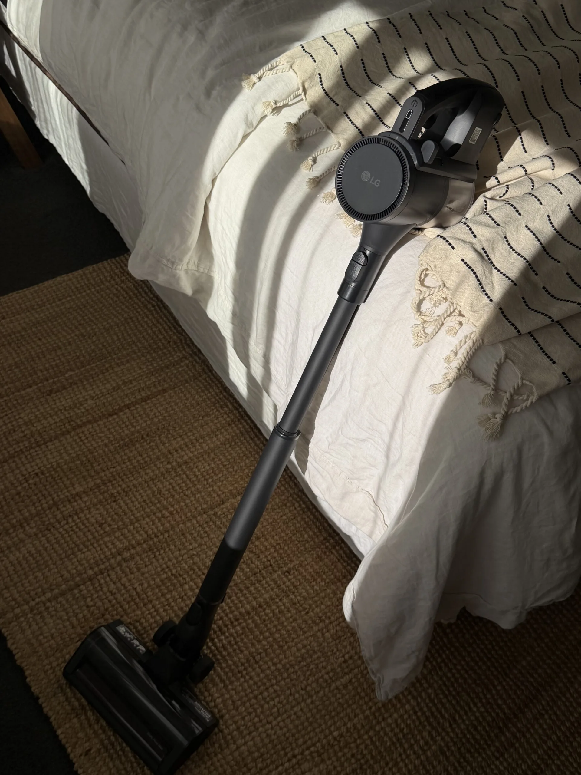 Cordless LG vacuum cleaner leaning against a bed with a woven rug in the foreground.