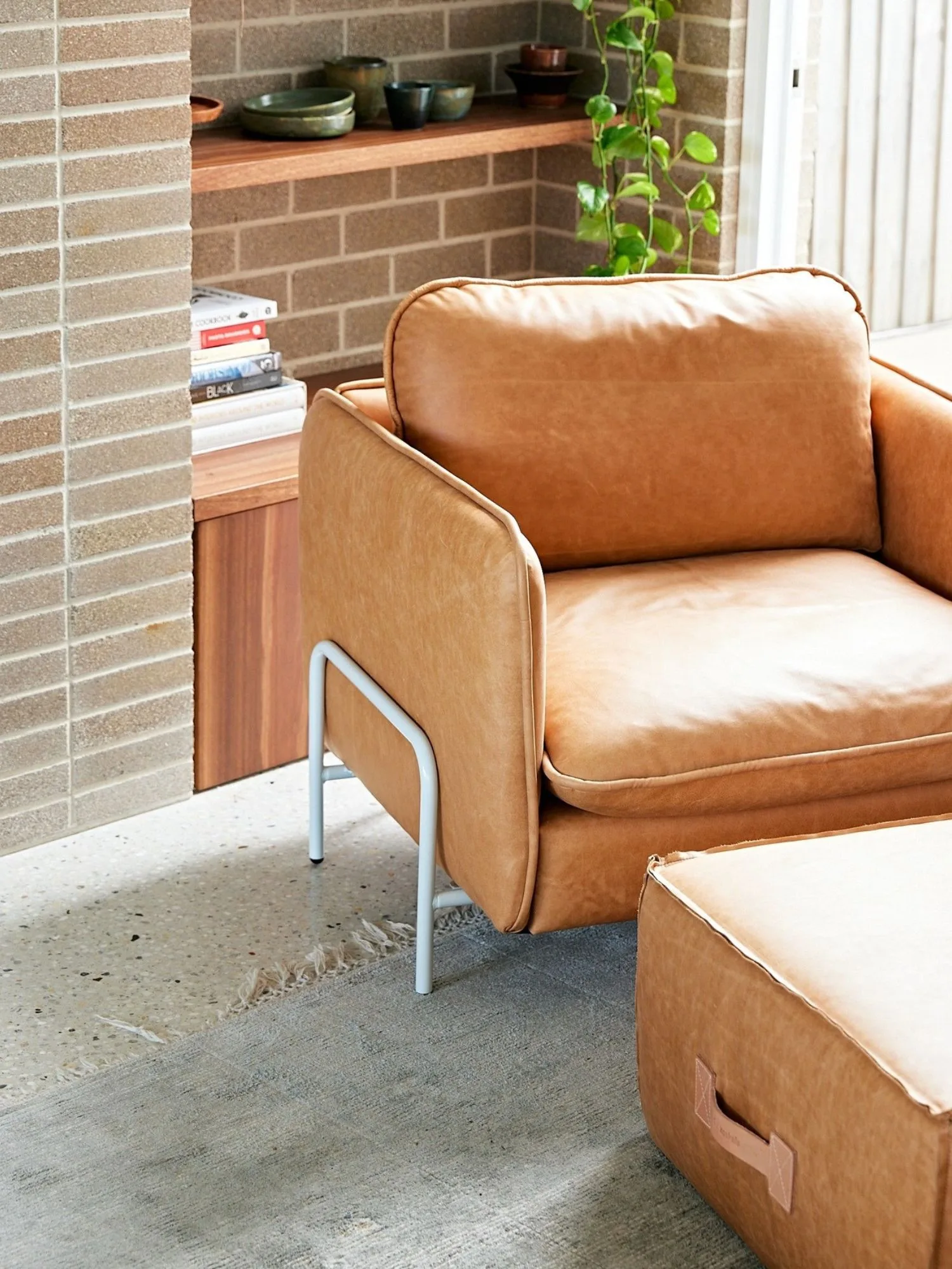 A modern tan leather armchair with a pale blue metal base, made in Australia by Koskela, sits in a living room with exposed brick wals