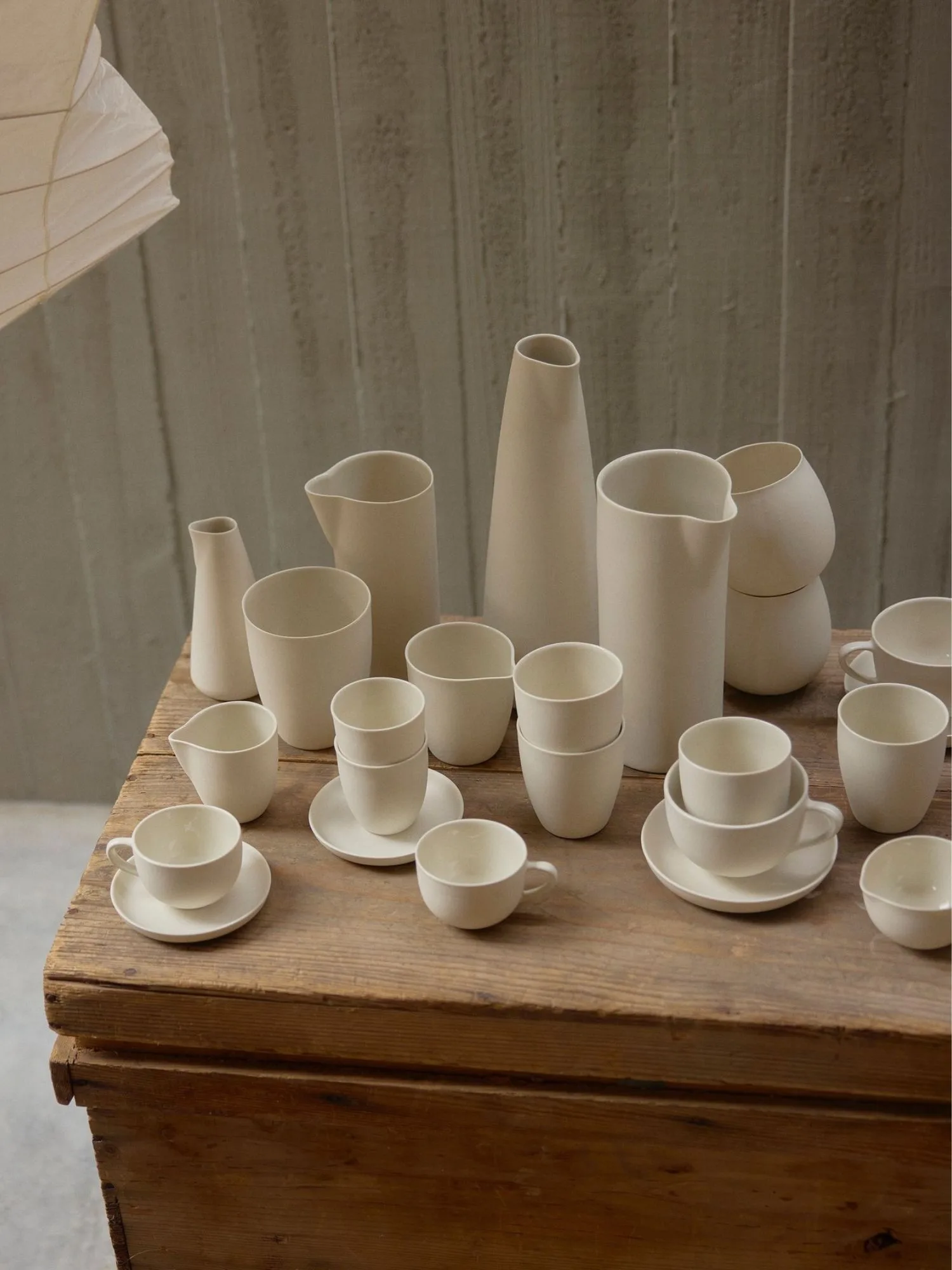 Dozens of different cream coloured, Australian-made ceramics by Mud Australia, including jugs, cups, vases and saucers, on a rustic raw timber surface.