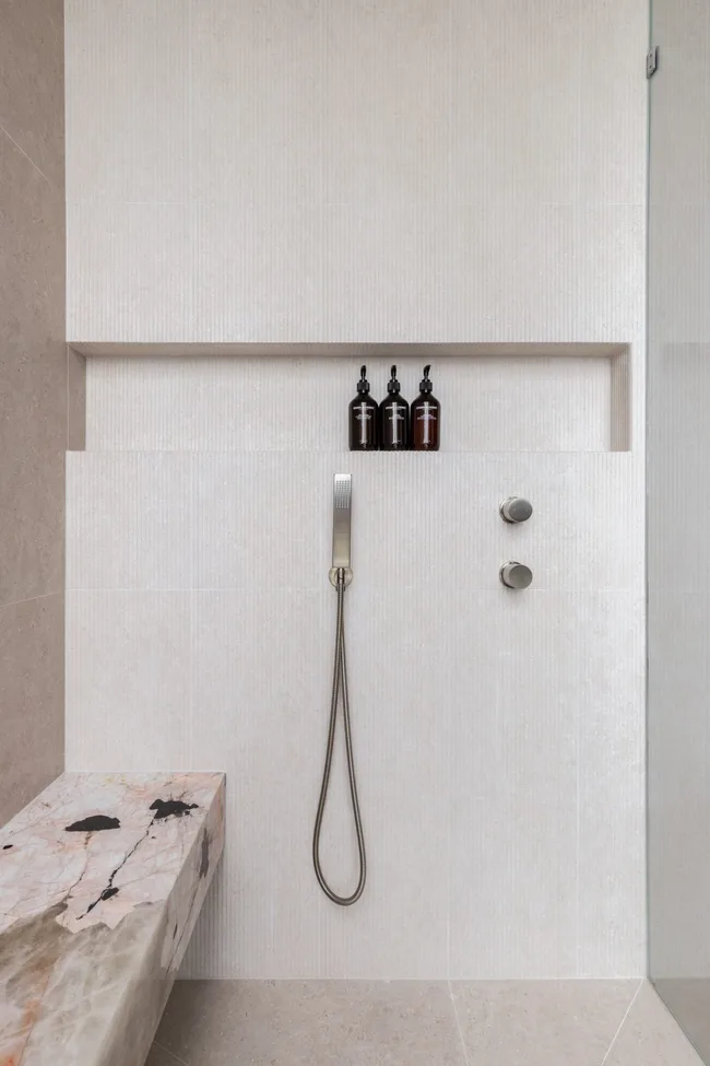 Modern shower with a handheld Chrome showerhead, warm white coloured tiles, marble shower bench, and three bottles in a recessed shower niche.