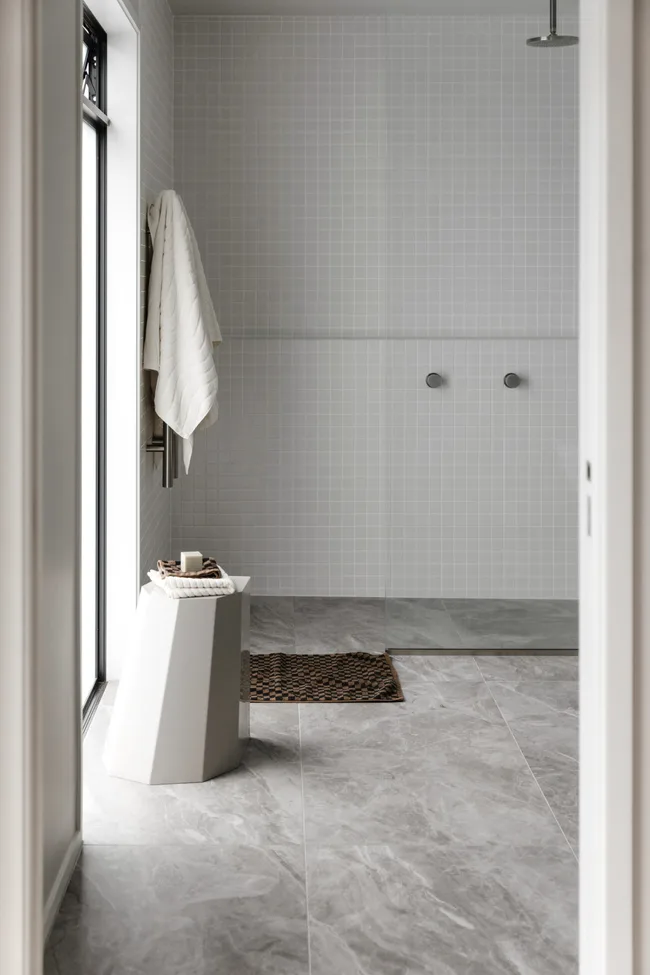 Modern bathroom with grey floor tiles, a white towel on a Chrome towel rack, and a geometric stool holding toiletries. The shower wall is white, with small square tiles, and a built-in shower ledge running the length of the wall.