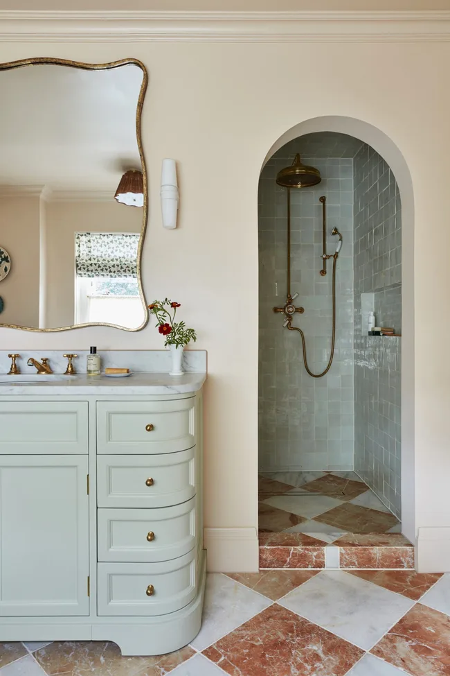 Elegant bathroom with light green vanity, an arched doorway leading to a shower with brass fixtures, built-in shower niche and glossy tiles. The vanity has a marble countertop, and the room features a patterned tile floor.