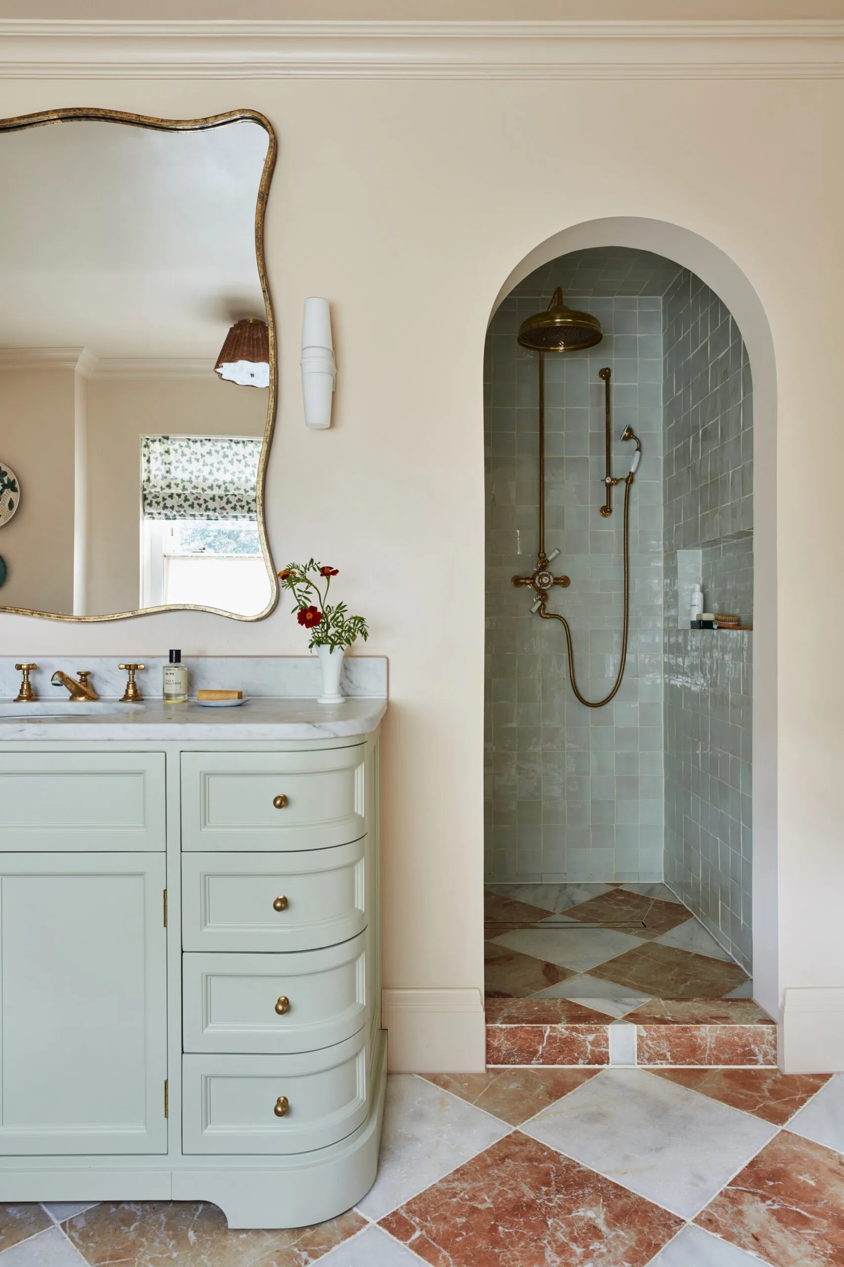 Elegant bathroom with light green vanity, an arched doorway leading to a shower with brass fixtures, built-in shower niche and glossy tiles. The vanity has a marble countertop, and the room features a patterned tile floor.