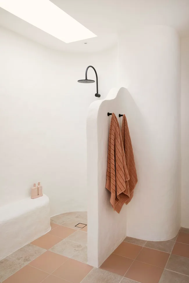 Bathroom with a European villa vibe. The walls are rendered and a curved white nib wall separates the shower from the rest of the room. There is also a curved shower bench. The tiled floor is orange and beige, with two orange towels on two shower hooks on the nib wall.