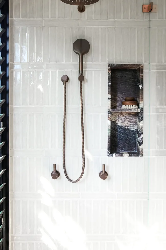 Modern shower with bronze fixtures on white tiled wall, with a shower niche made from black a dramatically veined and dark toned marble.