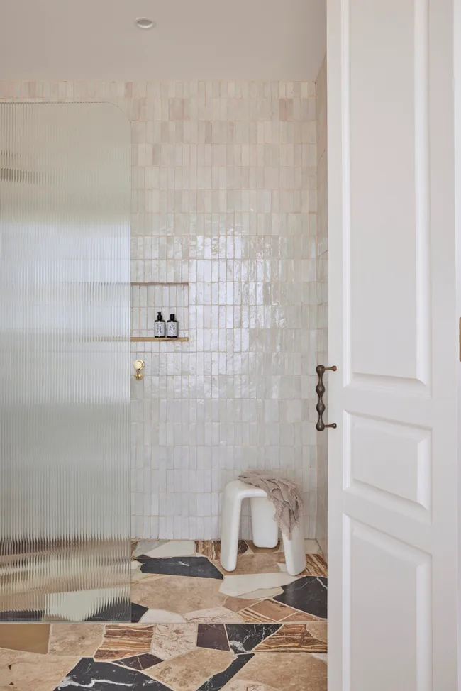 Bathroom with glossy tiled walls, a frosted glass shower screen, patterned tile floor, and a stool with a towel in the corner. The wall has a built-in shower niche.