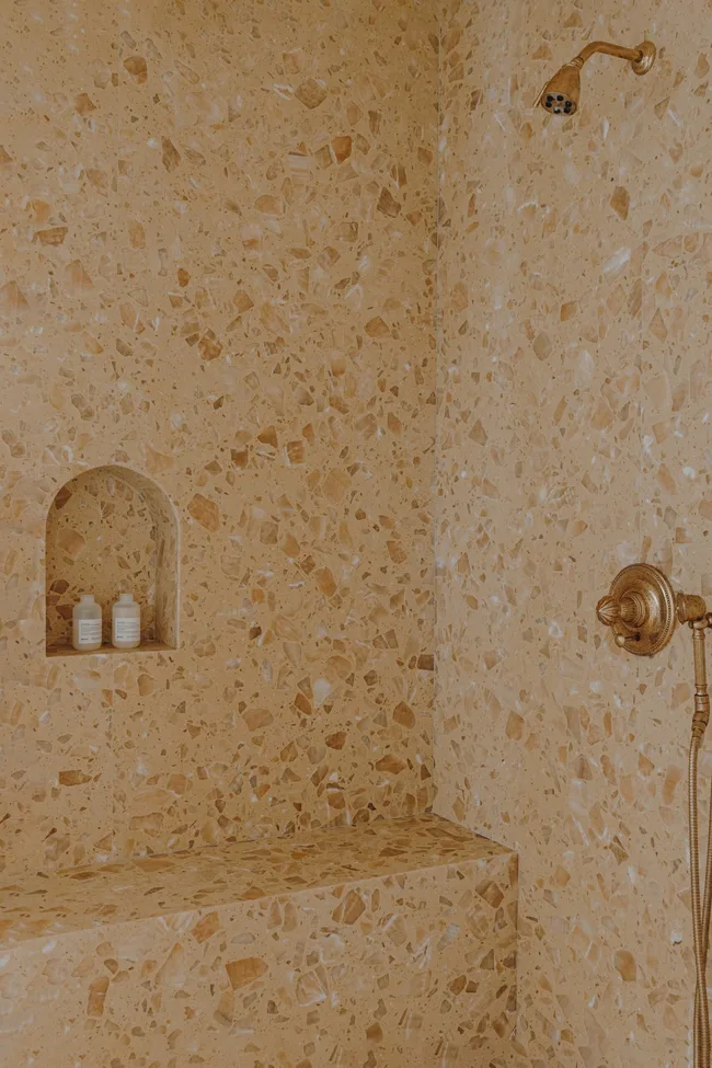 Shower with terrazzo walls, built-in arched shower niche with bottles, a shower bench built-in along one wall and brass fixtures.