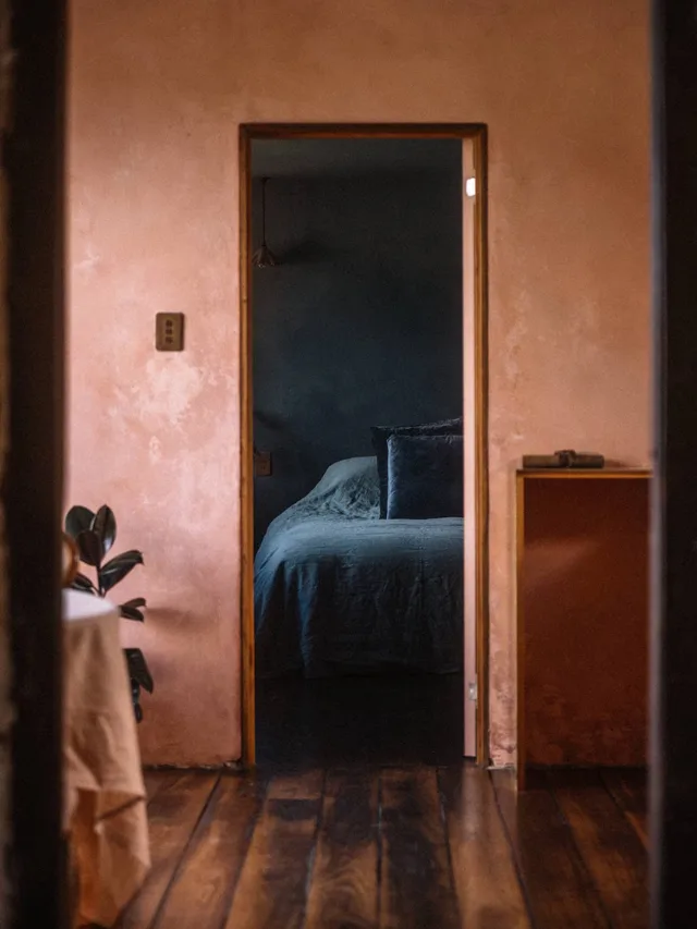 An image of the Historic Post House Airbnb in Carlton River, with limewashed walls in a warm shade of pink framing a dark blue bedroom with a plush bed