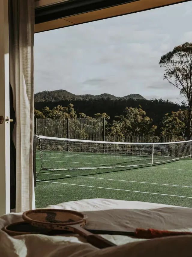 View of the tennis court from a bedroom at The Chalet & The Pavilion Airbnb in Spring Beach, Tasmania, with expansive bush in the background.