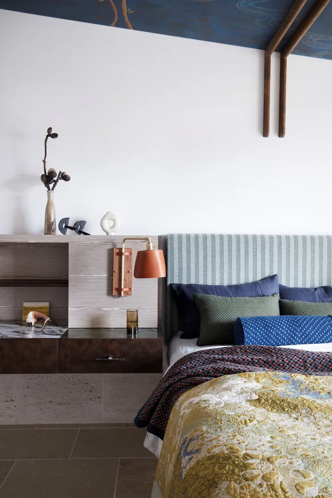 Cozy bedroom with a textured quilt, blue cushions, wall-mounted lamp, and decorative shelf above a bedside table.