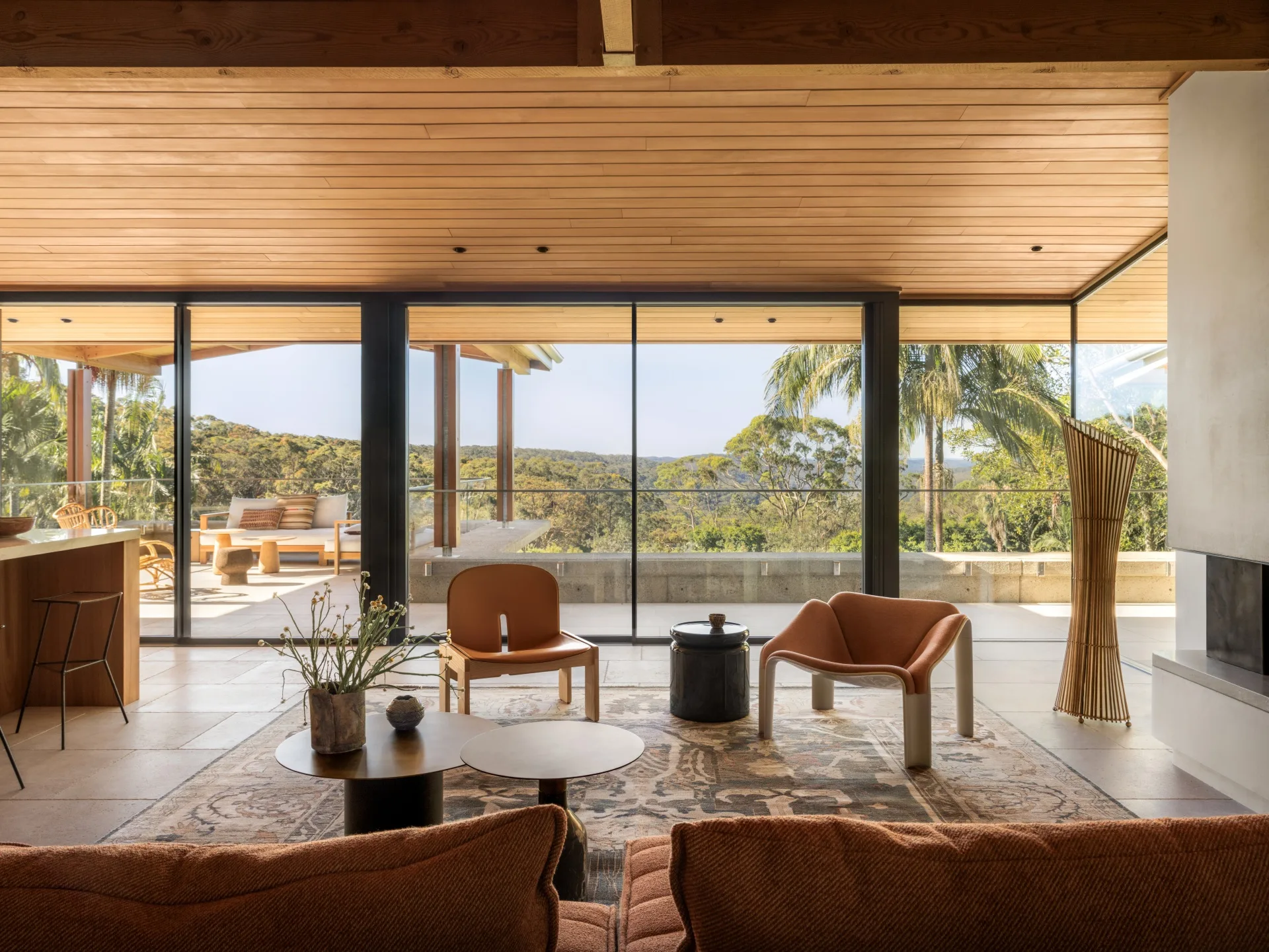 Modern living room with large windows, wooden ceiling, orange chairs, and a view of lush greenery outside.