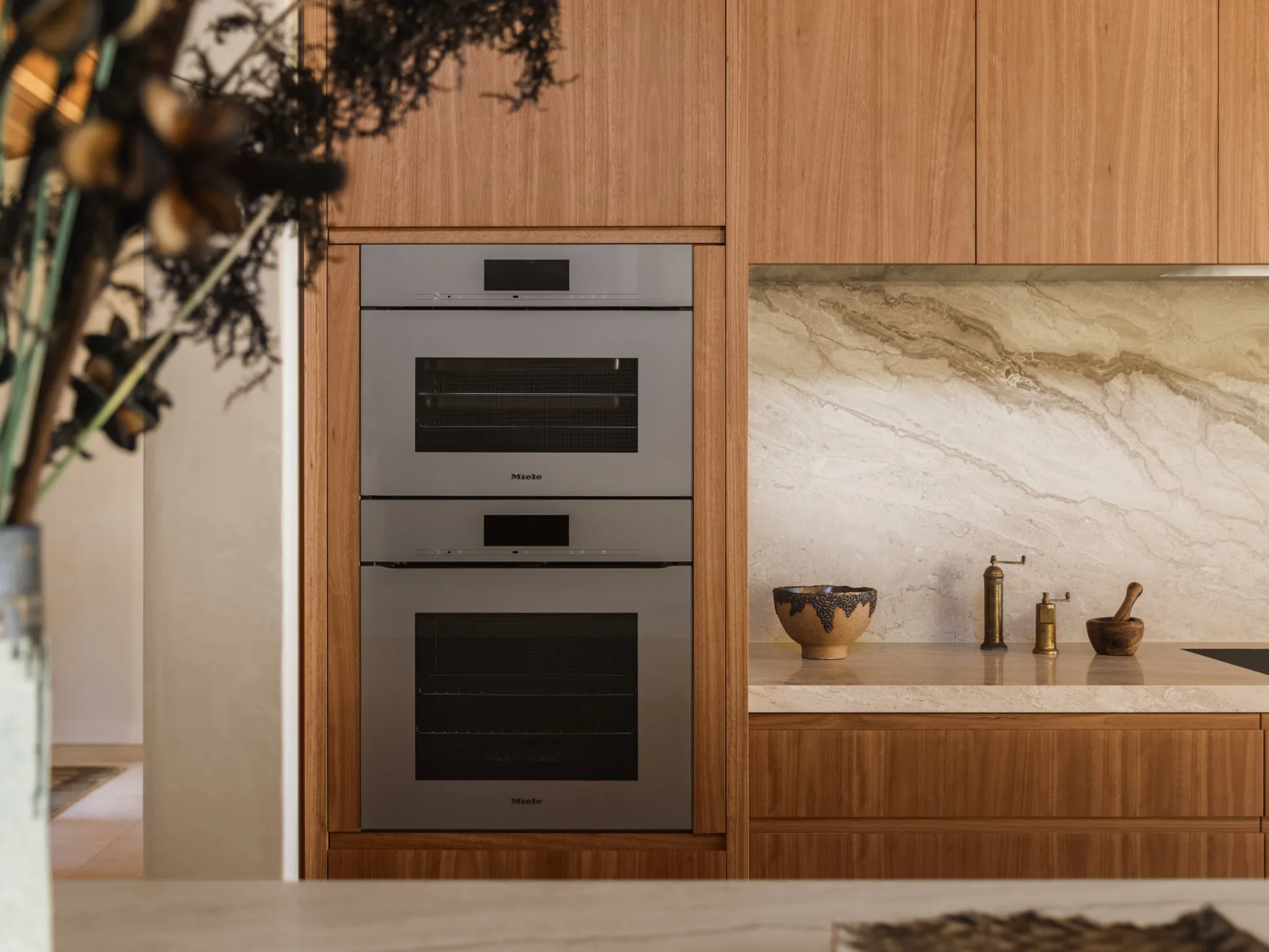 Built-in double oven in modern kitchen with wood cabinets, marble backsplash, and decorative items on the counter.