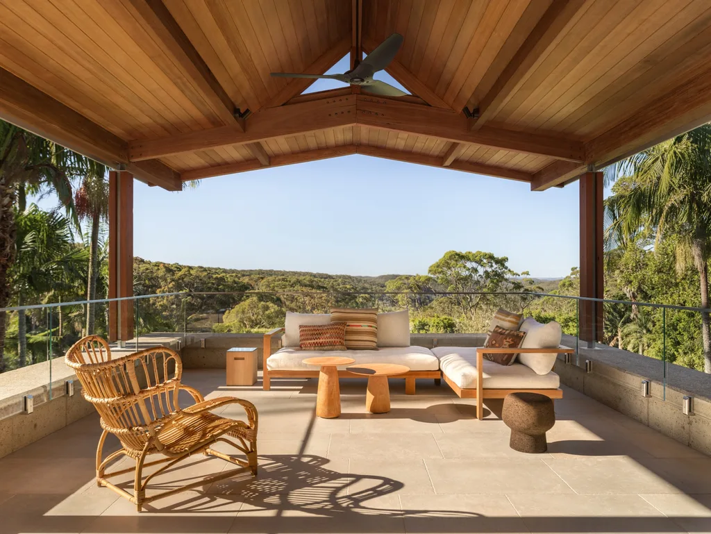 Covered outdoor patio with wooden ceiling, wicker chair, plush seating, and table, overlooking lush green landscape.