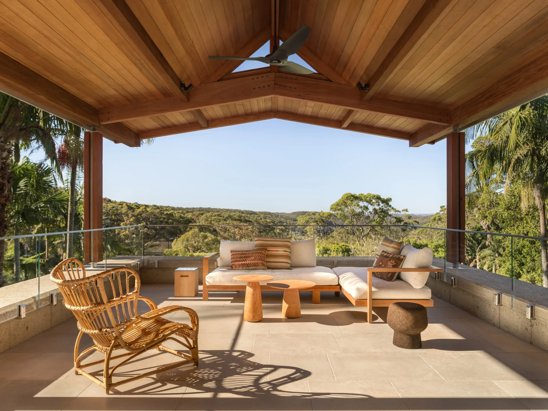 Covered outdoor patio with wooden ceiling, wicker chair, plush seating, and table, overlooking lush green landscape.