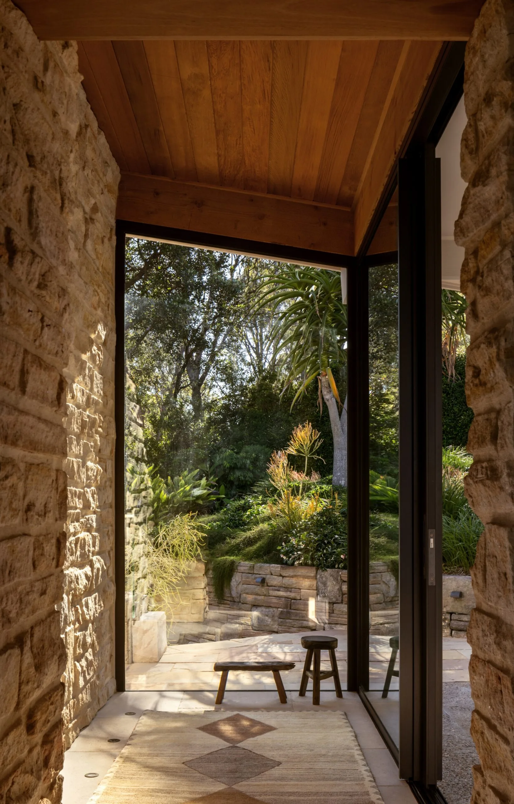 Covered stone patio with a view of a lush garden, featuring a small round table and stool against natural stone walls.