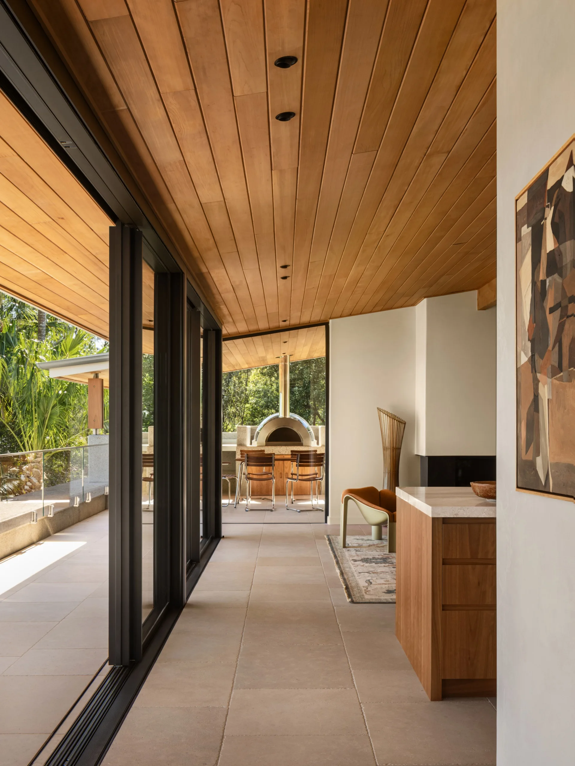 Modern interior with wooden ceiling, large glass doors, and outdoor pizza oven. Harmonious blend of indoor and outdoor spaces.