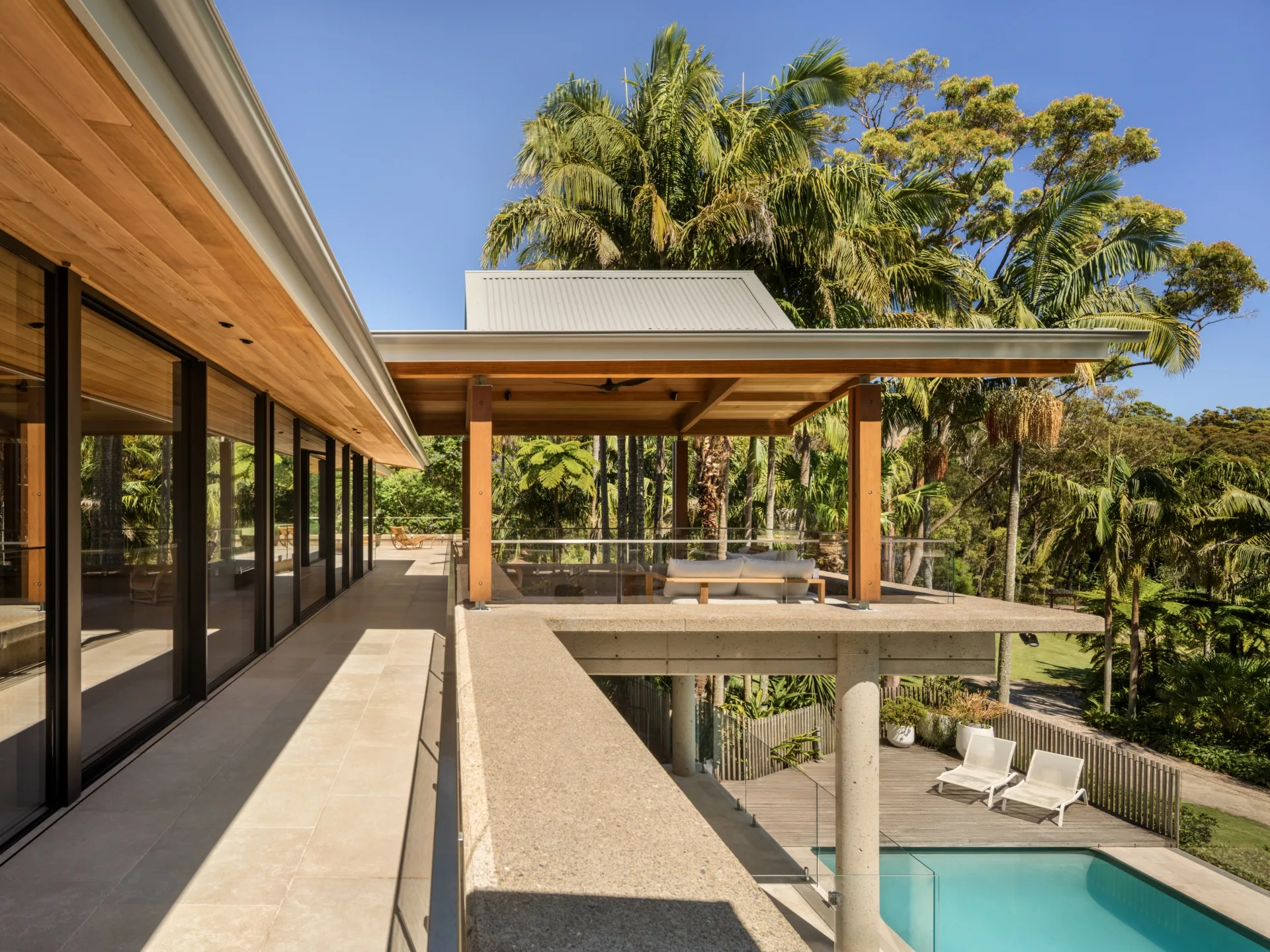 Modern house with a terrace overlooking a pool, surrounded by lush palm trees and tall windows reflecting the sky.