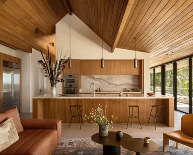 Modern kitchen with wooden ceiling, a marble island, brown sofa, decorative vase, and glass doors leading outside.