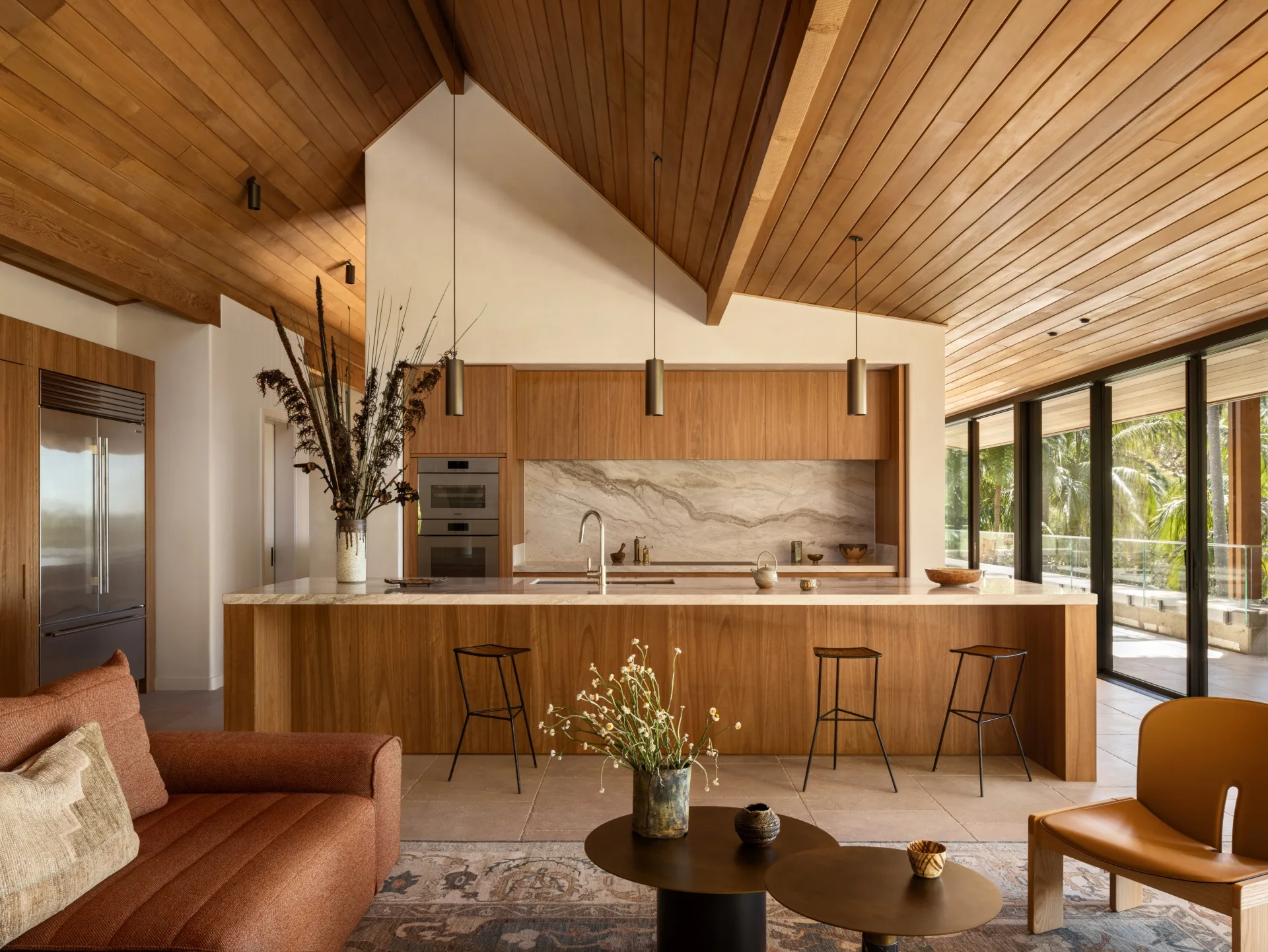 Modern kitchen with wooden ceiling, a marble island, brown sofa, decorative vase, and glass doors leading outside.