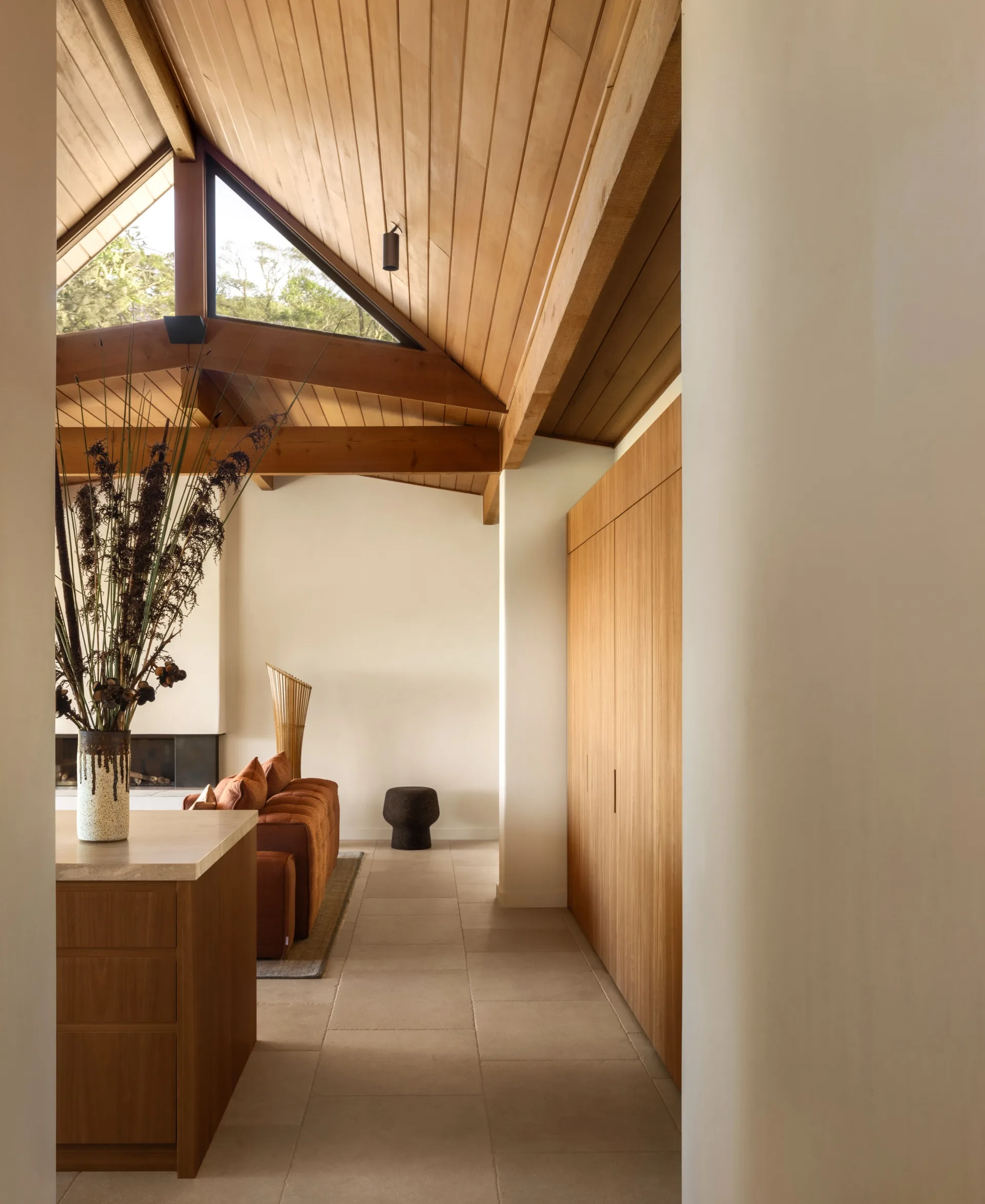 Modern living room with wooden ceiling, brown sofa, large vase on counter, and natural light from a triangular window.