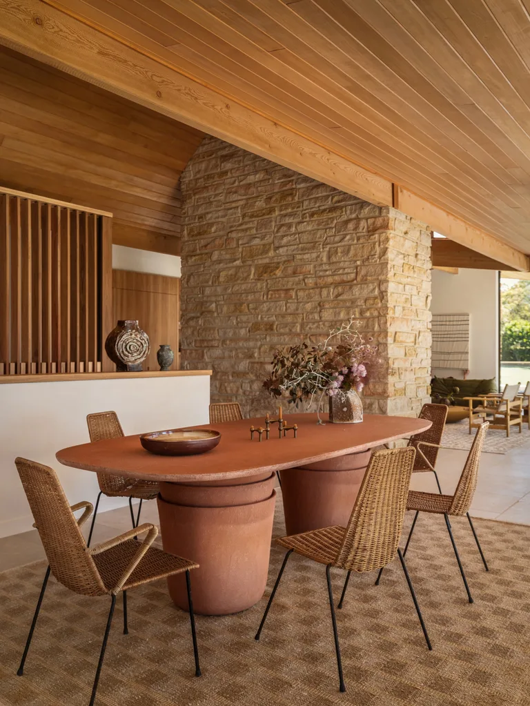 Dining area with terracotta table, wicker chairs, stone wall, wooden ceiling, and decor pieces on a checkered rug.
