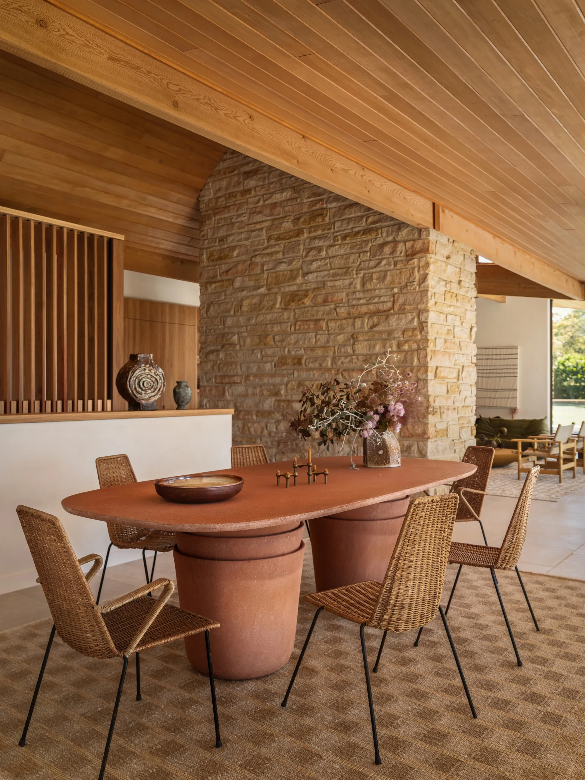 Dining area with terracotta table, wicker chairs, stone wall, wooden ceiling, and decor pieces on a checkered rug.