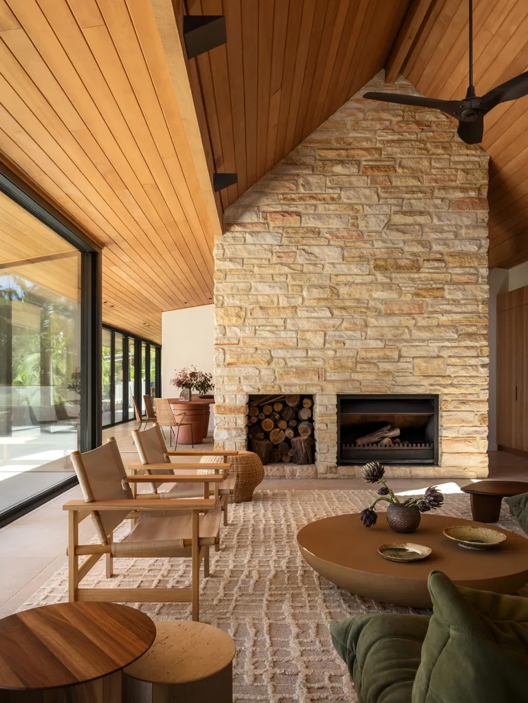 Warm, modern living room with wooden ceiling, stone fireplace, beige chairs, and large windows.