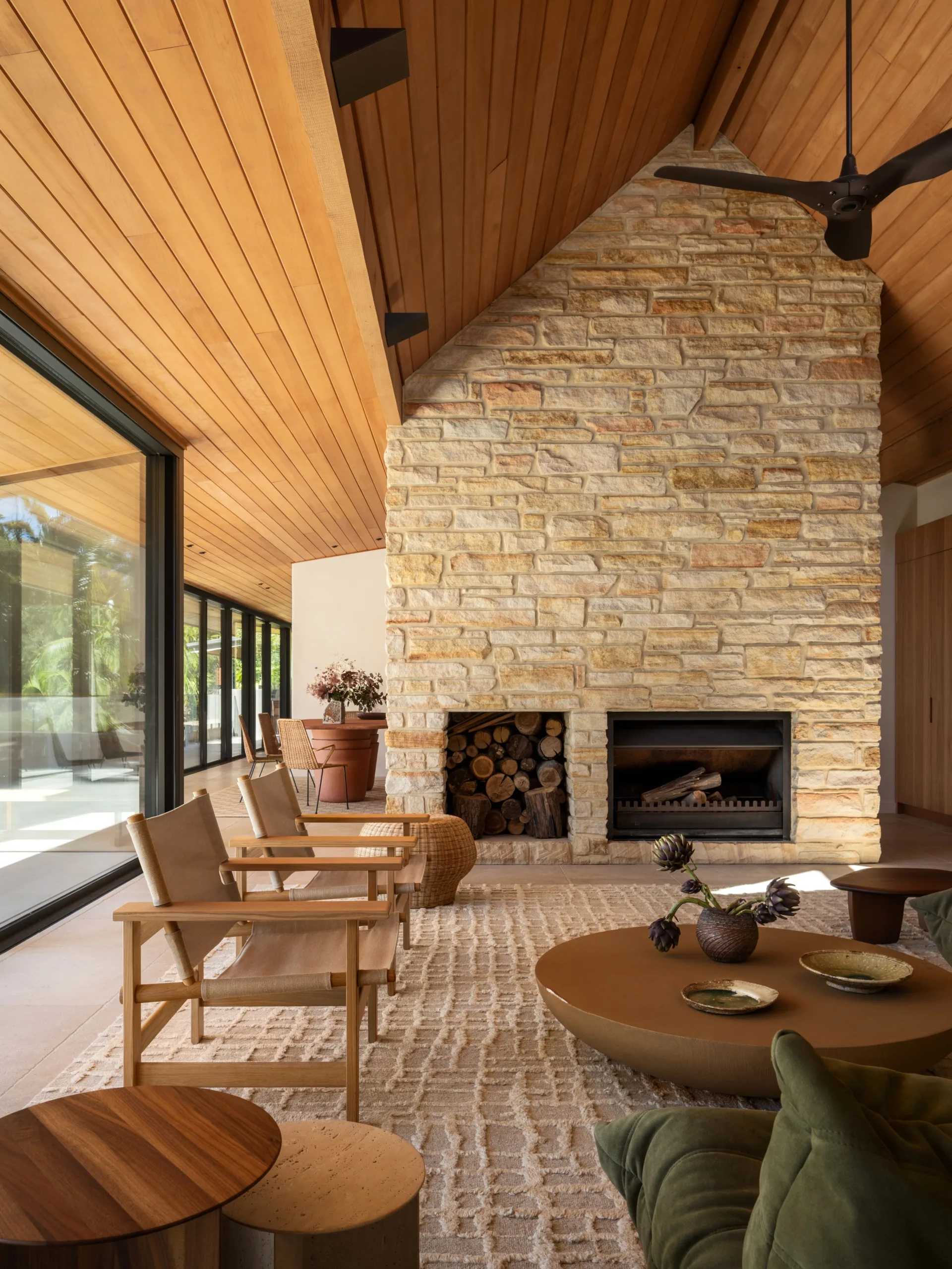Warm, modern living room with wooden ceiling, stone fireplace, beige chairs, and large windows.