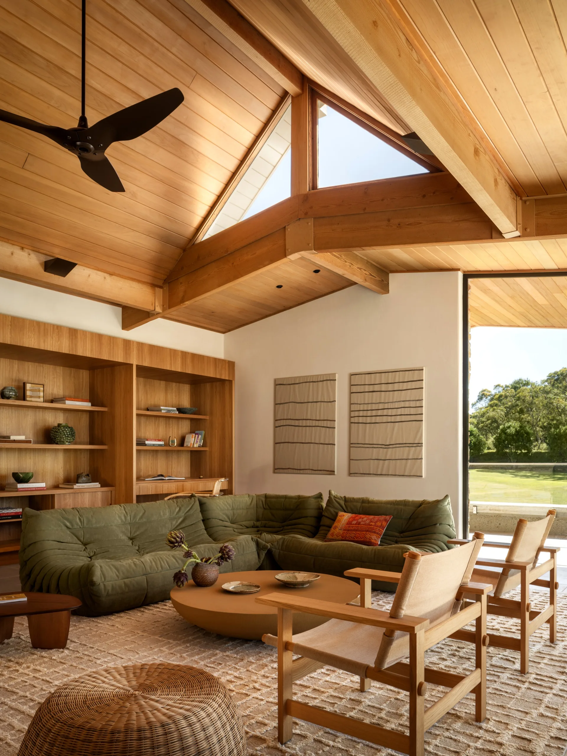 "Living room with wooden ceiling, green sectional sofa, round table, wicker stools, and chairs by a large window."