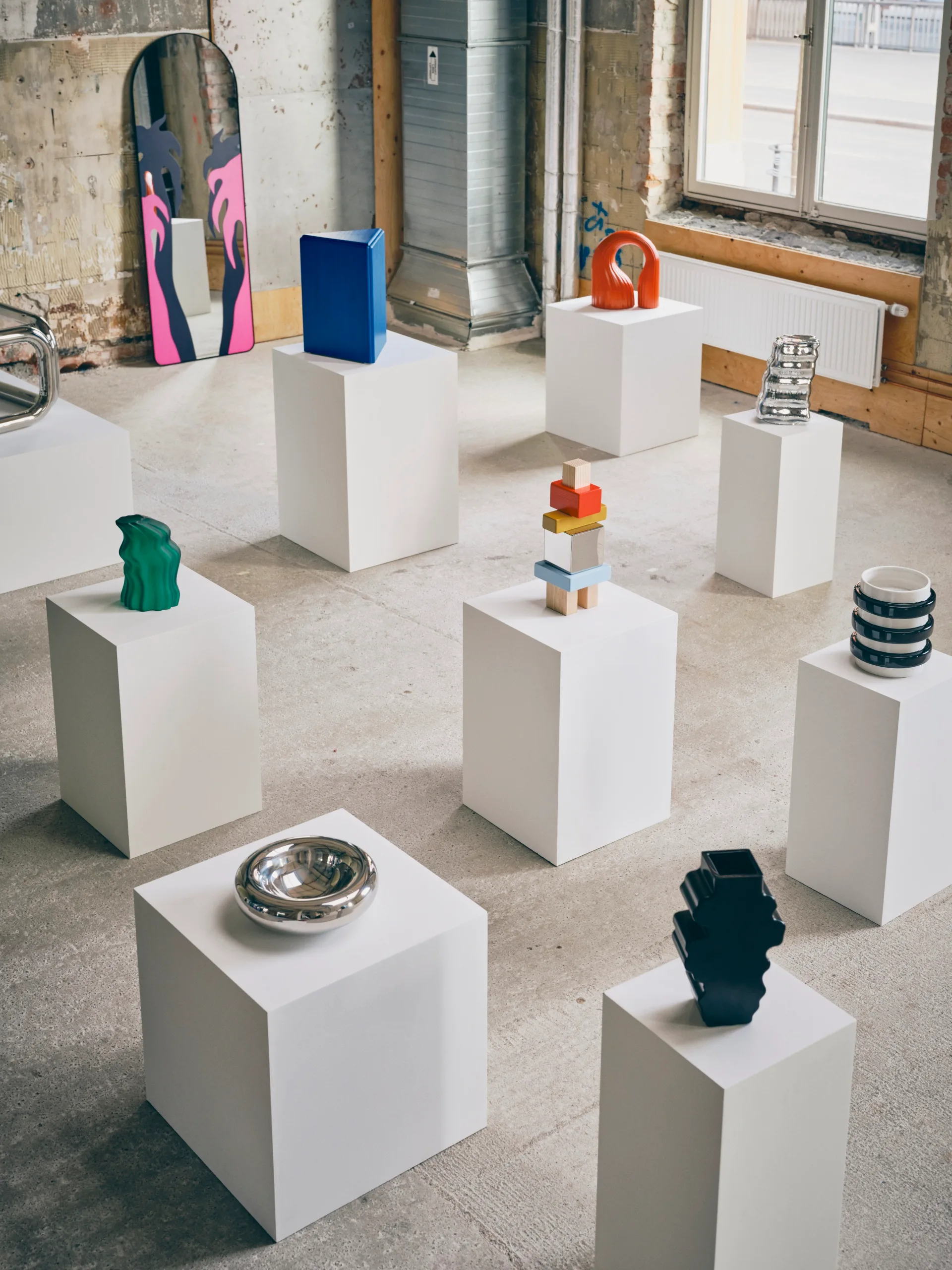 Colourful sculptures and decorative objects from the IKEA KONSTRUNDA collection displayed on white plinths in an industrial gallery space.