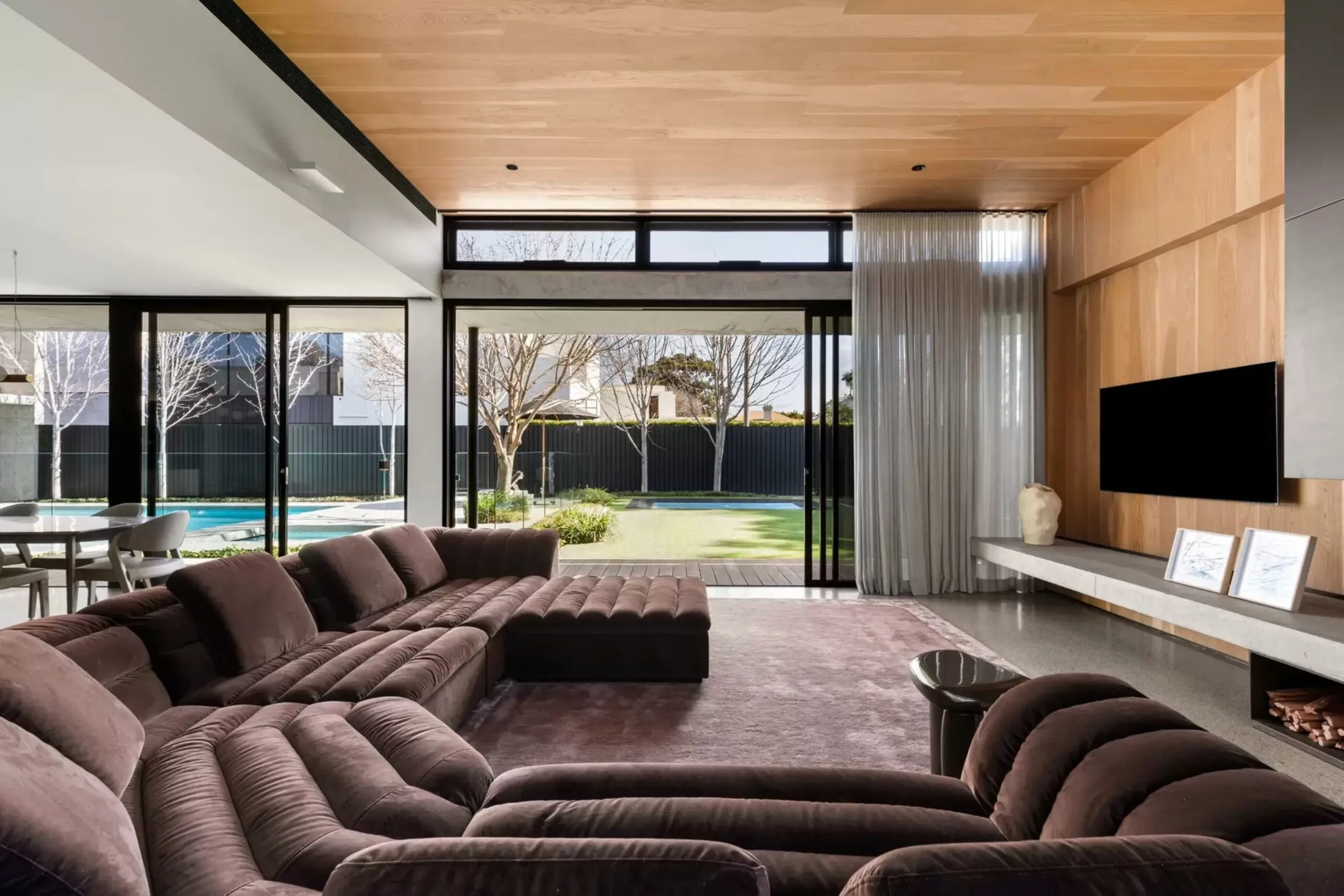 Living room with brown modular sofas, timber wall panelling and floor-to-ceiling windows overlooking the pool.