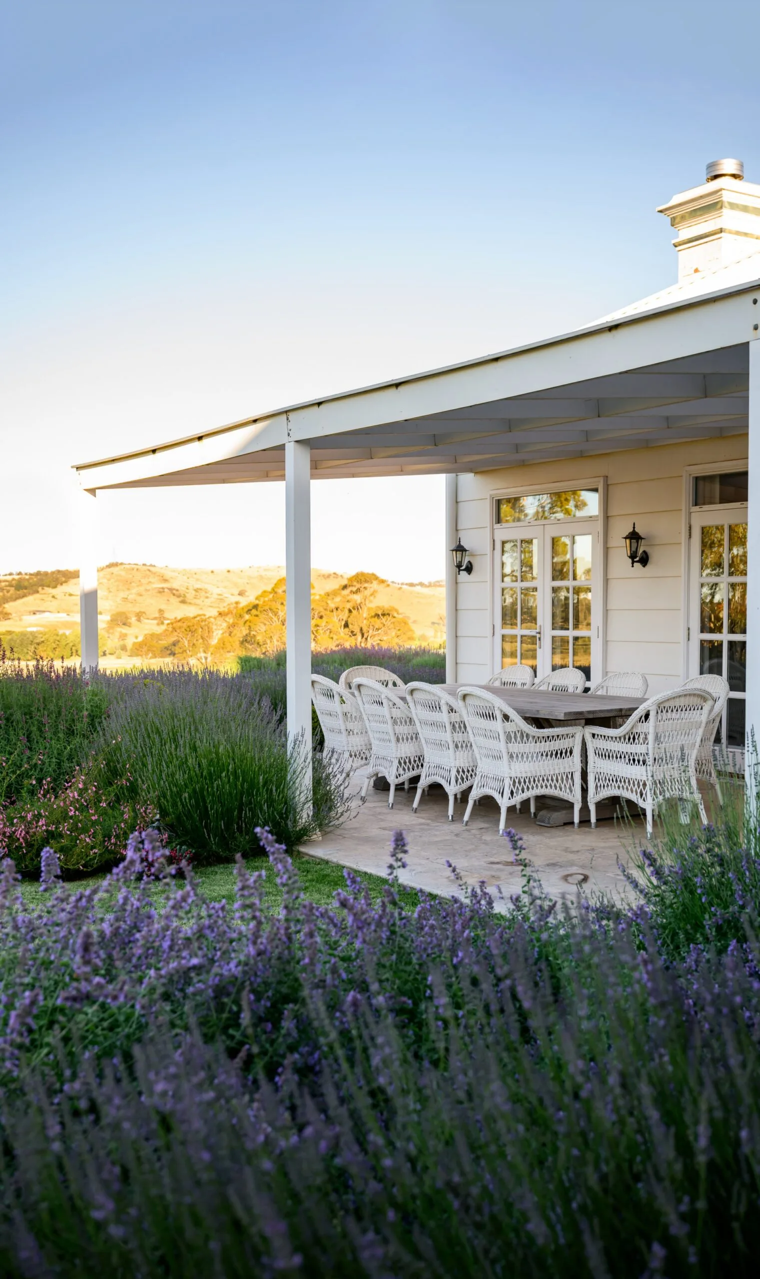 White house in country Australia with a veranda, alfresco dining area including white dining chairs, and a European-style garden in the foreground under a clear blue sky. Lavender and star jasmine are visible in the foreground.
