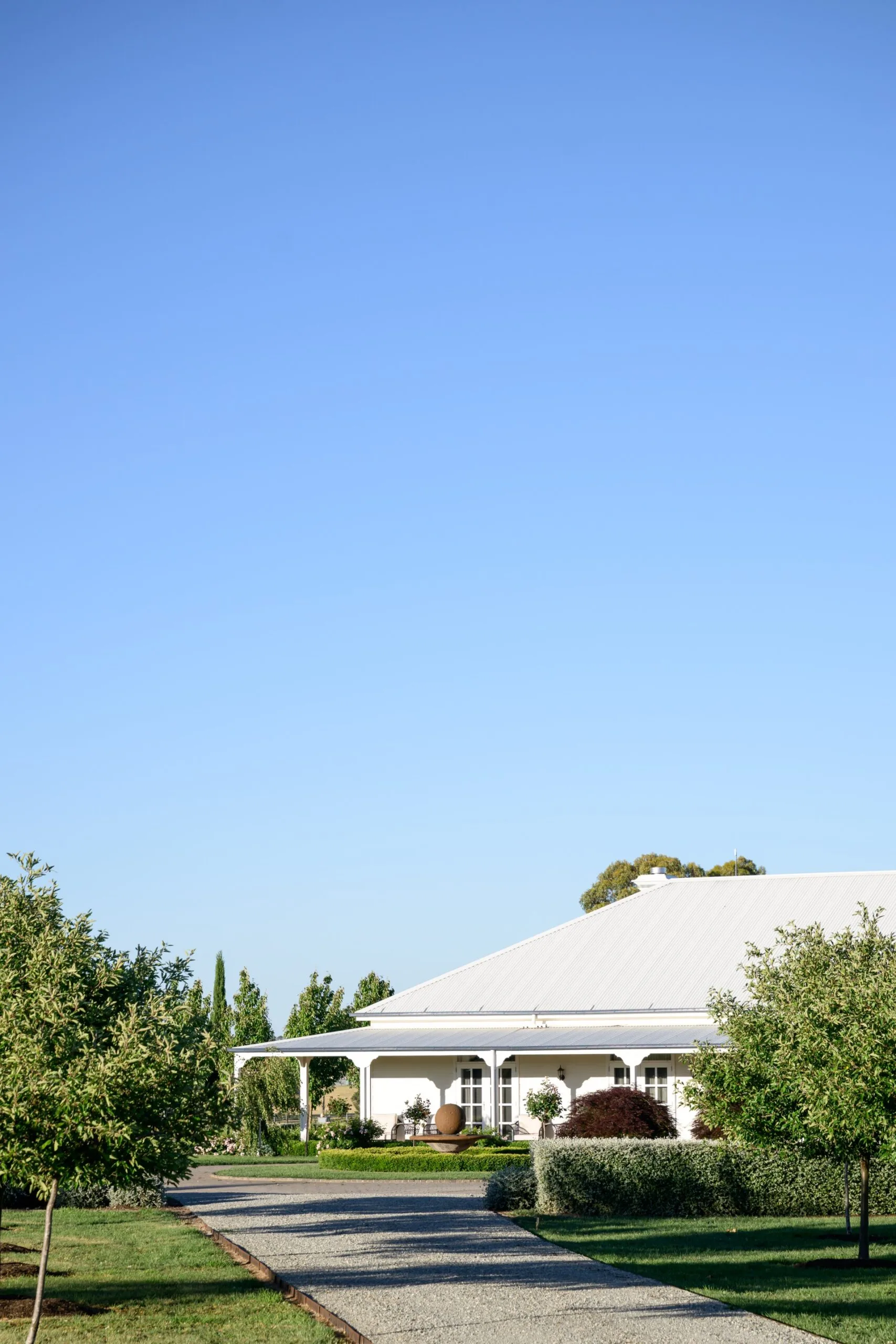 A classic Australian country home with a white facade. A driveway leads up to the home, with hedges and trees on either side. A small fountain is in the middle of the circular driveway, surrounded by hedges. The sky is blue and cloudless.
