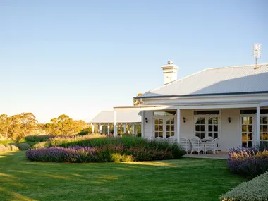 White house in country Australia with a veranda, alfresco dining area including white dining chairs, and a European-style garden in the foreground under a clear blue sky. Lavender and star jasmine are visible in the foreground.