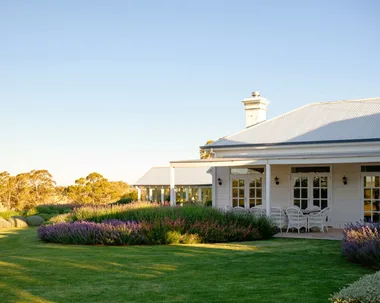 White house in country Australia with a veranda, alfresco dining area including white dining chairs, and a European-style garden in the foreground under a clear blue sky. Lavender and star jasmine are visible in the foreground.