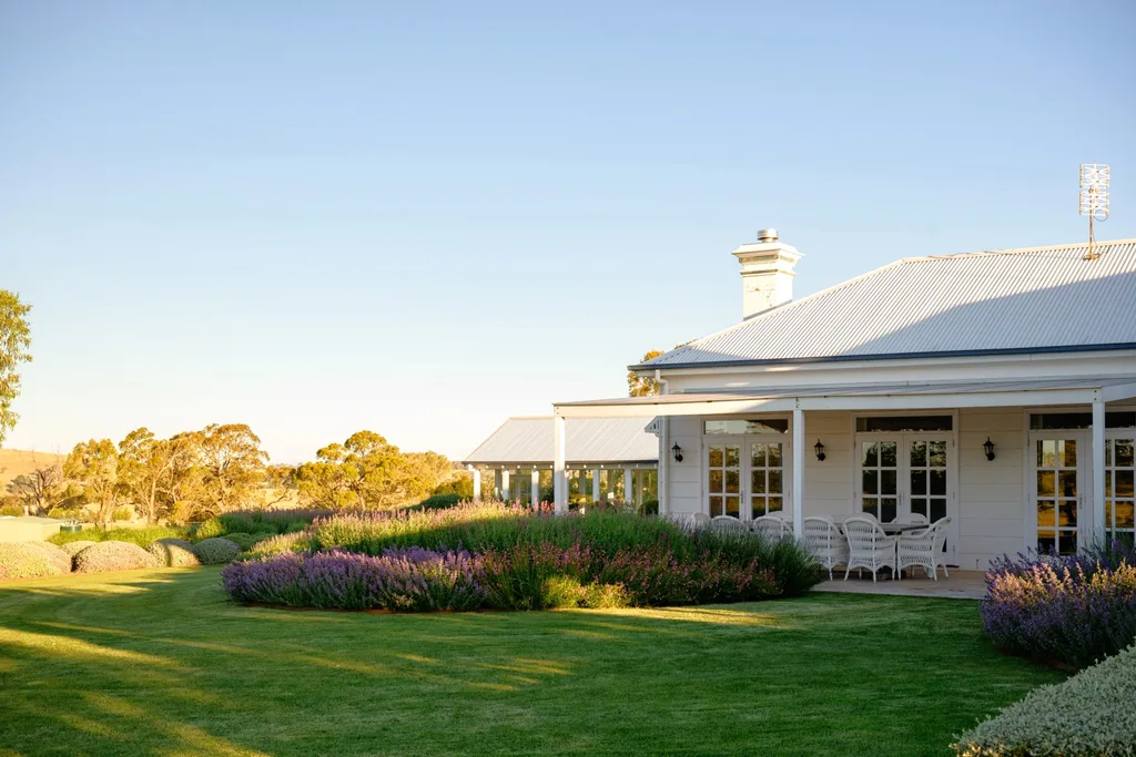 White house in country Australia with a veranda, alfresco dining area including white dining chairs, and a European-style garden in the foreground under a clear blue sky. Lavender and star jasmine are visible in the foreground.