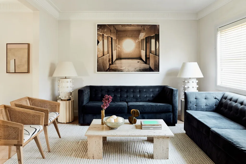 A living room with white walls a deep blue velvet sofa, white accent table lamps and a brown artwork