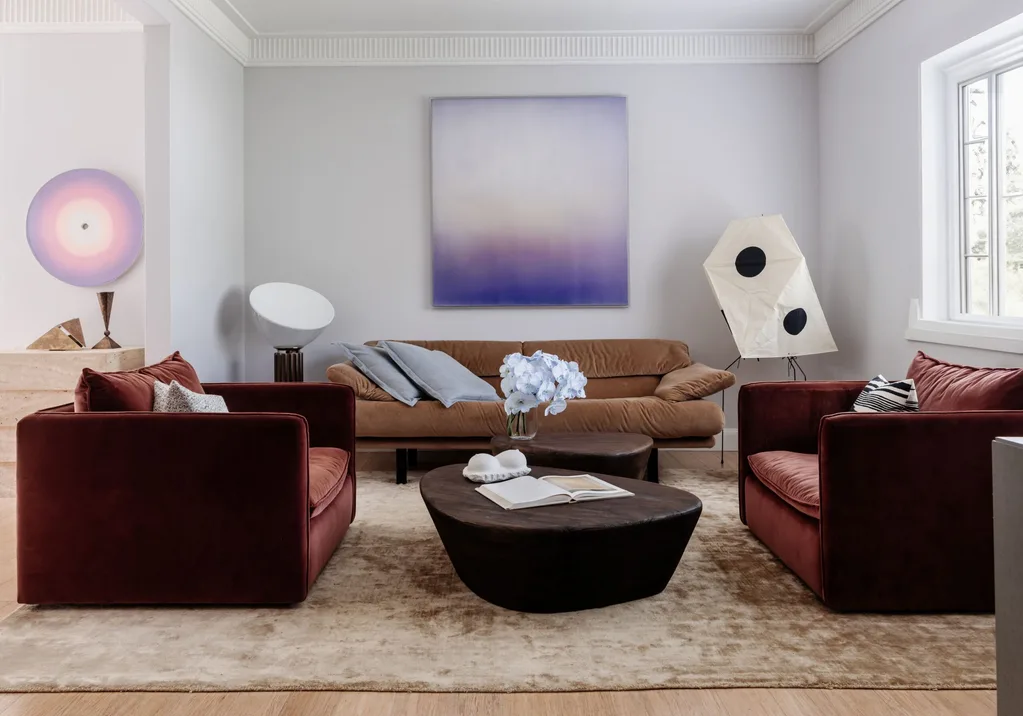 A living room with light mauva walls, a purple artwork, brown couch, red armchairs and brown coffee table