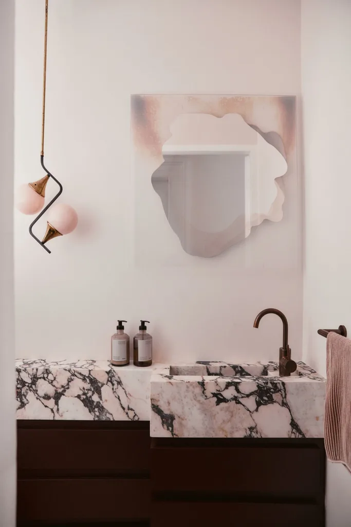 A bathroom vanity with marble countertops, a brass tap and a creative blush toned, frameless mirror