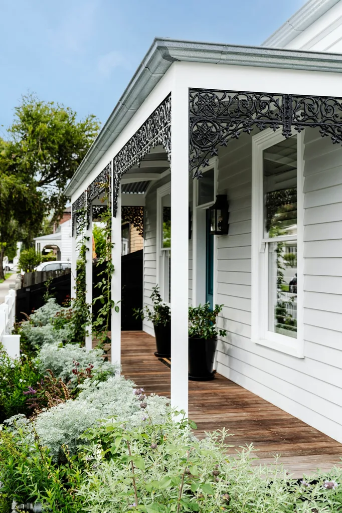 Front porch of a white house with ornate black ironwork, wooden flooring, and lush green garden.