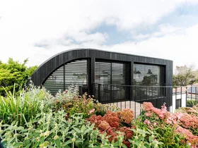 Modern black house with large windows, surrounded by a lush garden with various plants and flowers.