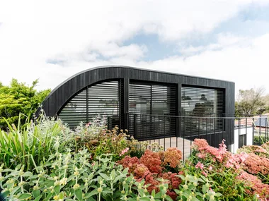 Modern black house with large windows, surrounded by a lush garden with various plants and flowers.