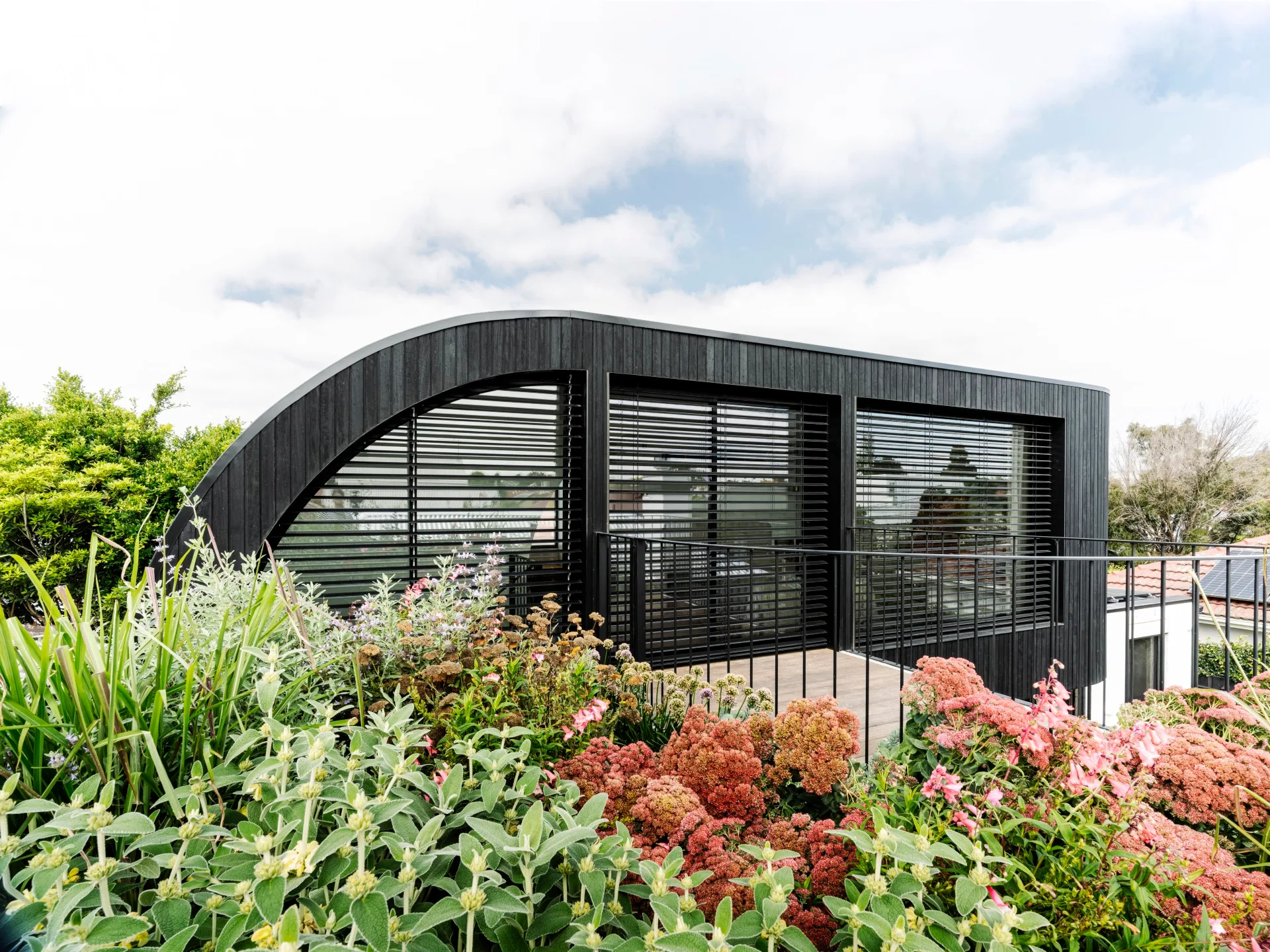 Modern black house with large windows, surrounded by a lush garden with various plants and flowers.