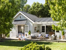 White cottage with children sitting on the porch, surrounded by trees and greenery under a clear sky.