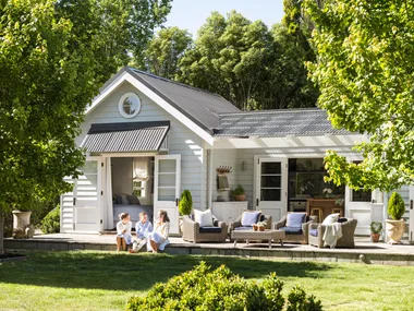 White cottage with children sitting on the porch, surrounded by trees and greenery under a clear sky.