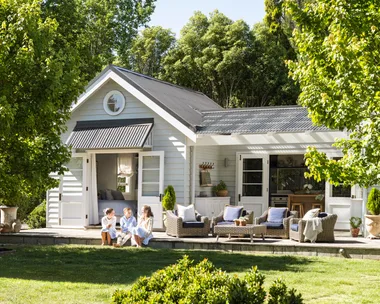 White cottage with children sitting on the porch, surrounded by trees and greenery under a clear sky.