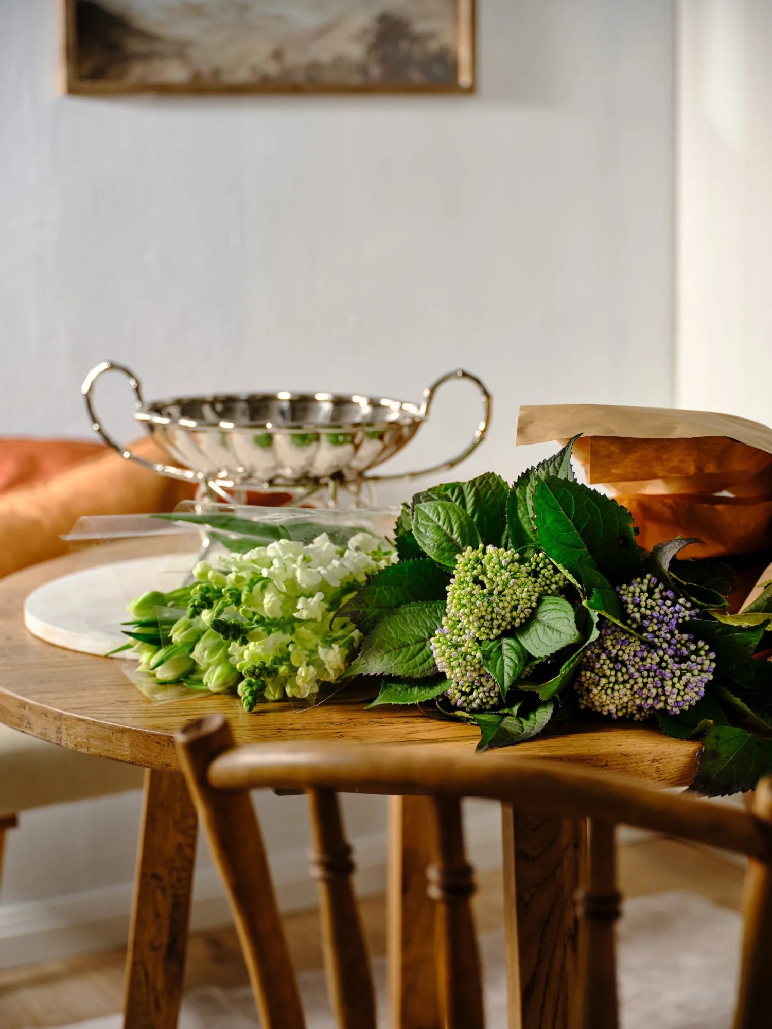Silver bowl and green floral bouquet on wooden table in a cozy room setting.