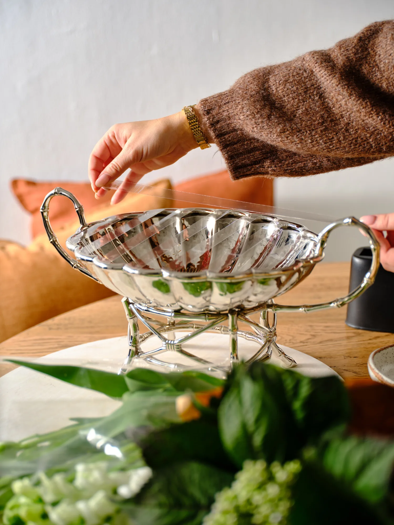 A hand placing an item into a silver bowl with handles on a wooden table, surrounded by greenery.
