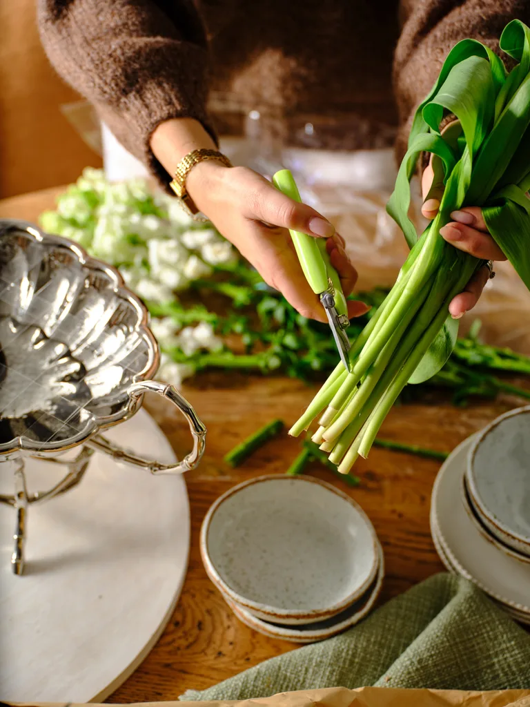 A person trims green stems with scissors at a wooden table, surrounded by plates and green leaves.